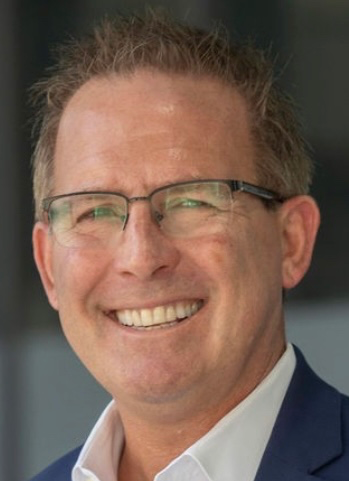 Man with glasses smiling, wearing a blue blazer and white shirt, outdoors.