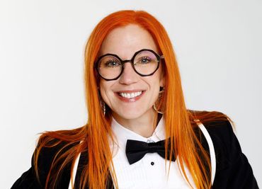 Woman with orange hair and glasses, wearing a bow tie and suspenders, smiling.