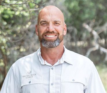 Bald man with a grey beard smiles, wearing a light blue collared shirt outdoors.