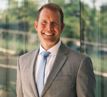 Man in gray suit and light blue tie smiles broadly in a modern setting.