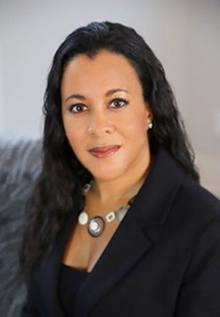 Woman with dark hair, wearing a black blazer and necklace, smiling, soft focus background.