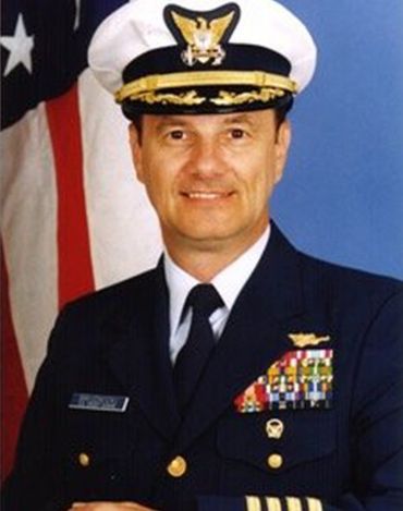 Man in U.S. Coast Guard uniform, wearing cap, facing camera, smiling slightly. American flag in background.