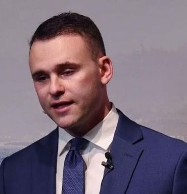 Man in a blue suit and tie speaking at a podium, set against a blurred background.