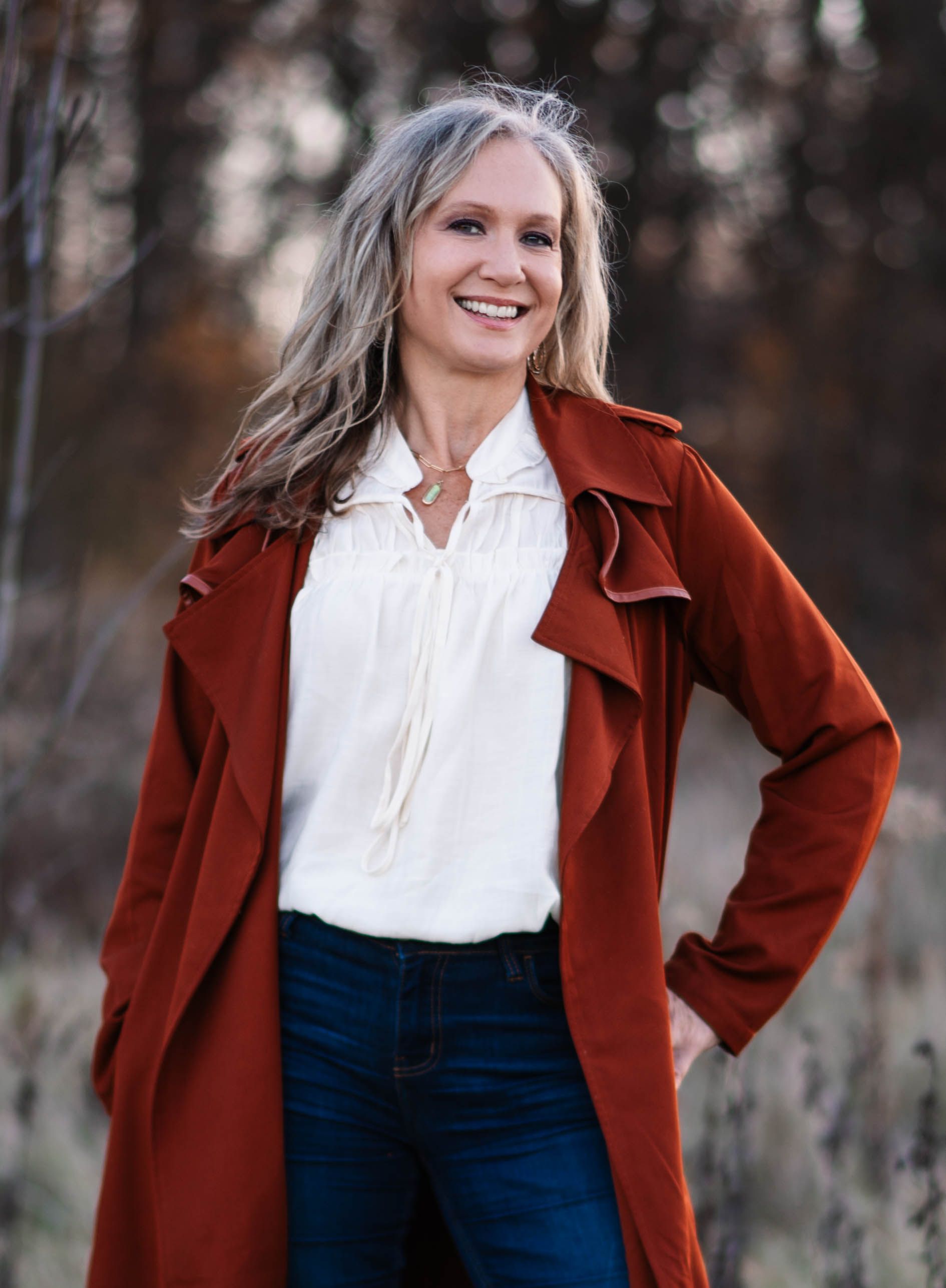 Woman in rust-colored coat smiles, standing outdoors with hand in pocket.