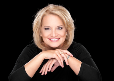 Woman with blonde bob smiles, resting arms on desk. Black background.