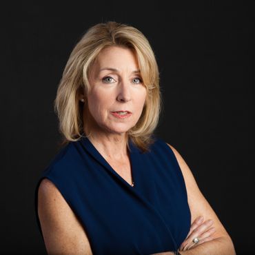 Woman with blonde hair, wearing a blue sleeveless top, arms crossed, looking at the camera against a black background.