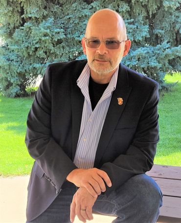Bald man in blazer, sunglasses, and jeans, kneeling outdoors in front of a tree.