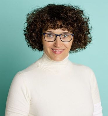 Woman with curly brown hair wearing glasses and a white turtleneck smiles, turquoise background.