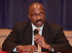 Man in suit speaking into a microphone at a table. He wears glasses, a tie, and a watch.