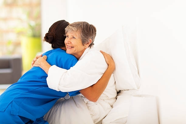 A healthcare worker in blue scrubs hugs an older person in a white robe, both smiling. Bright setting.