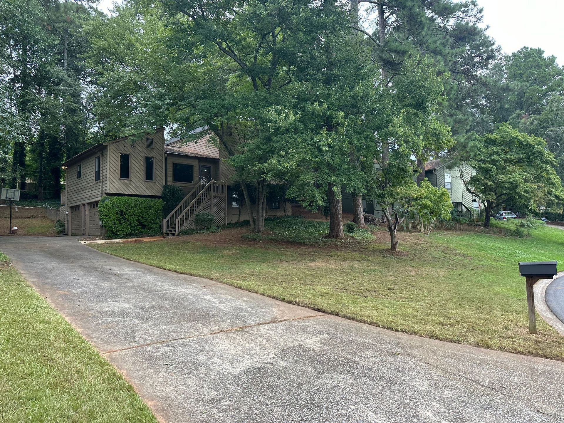 House with a driveway, trees, and mailbox on a grassy lawn. The house has a wooden deck and siding.