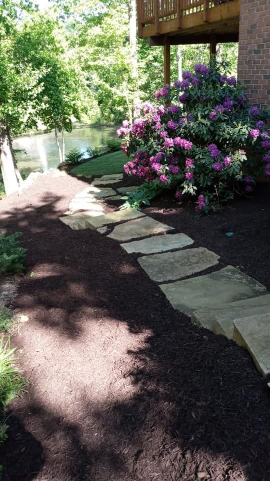 Stone path through mulch, past a blooming purple rhododendron bush, leading to a wooden deck.