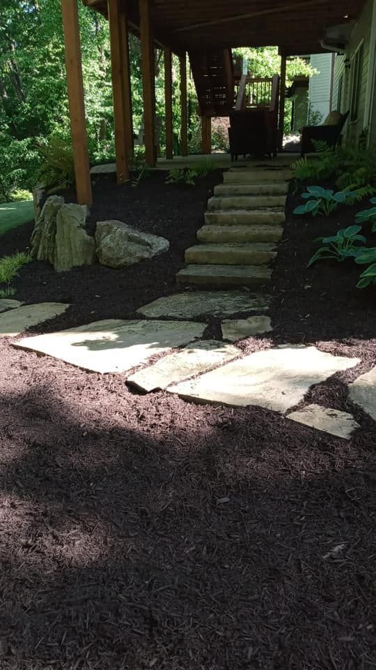 Stone path and stairs leading to a covered porch; dark mulch, lush greenery, and dappled sunlight.