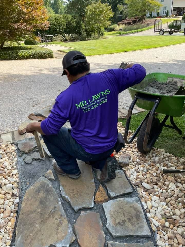 Man in purple shirt installing stone walkway, using a trowel and hammer. Green wheelbarrow nearby.