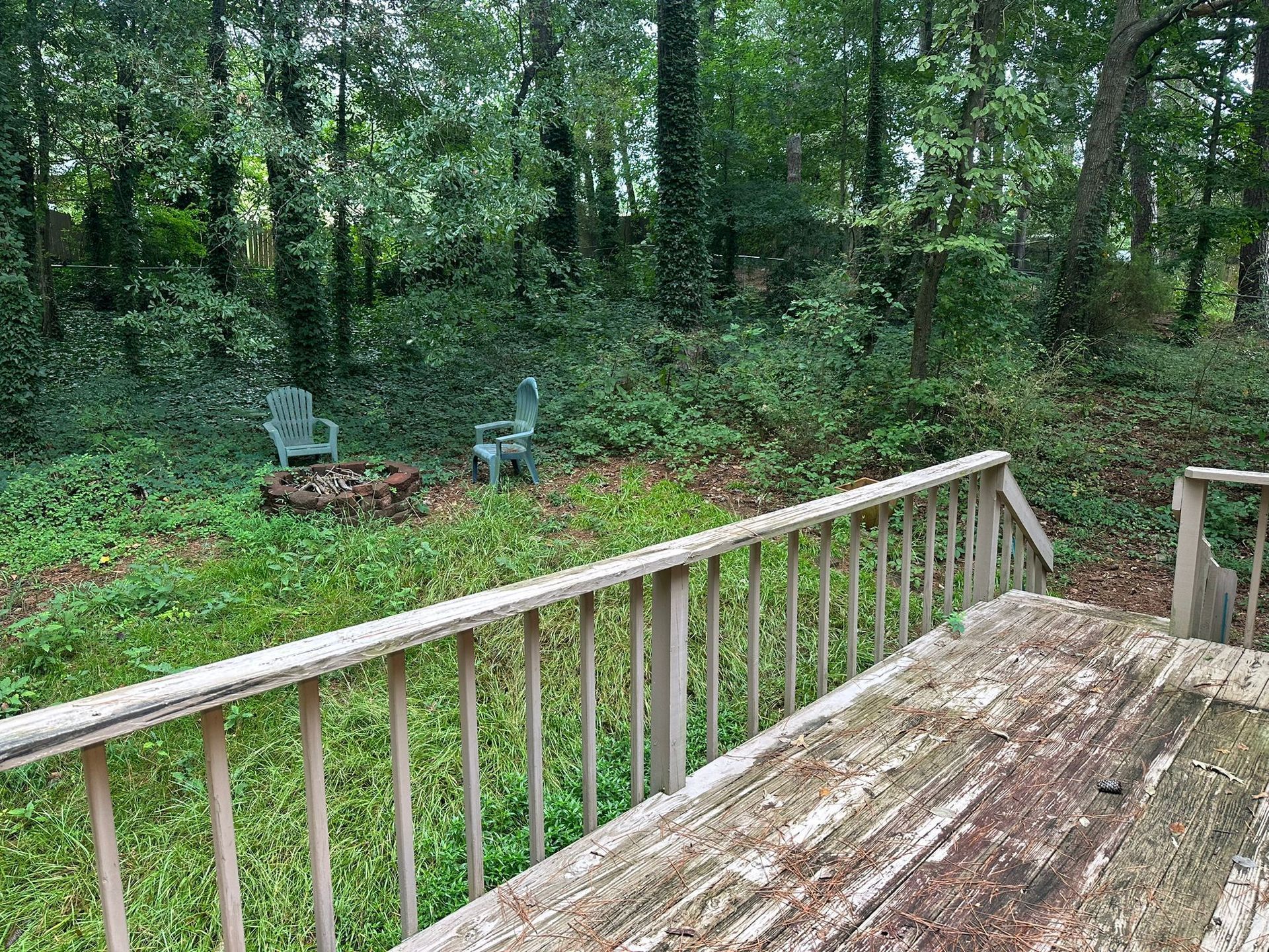 View from weathered deck overlooking a backyard with two chairs, a fire pit, and lush greenery.