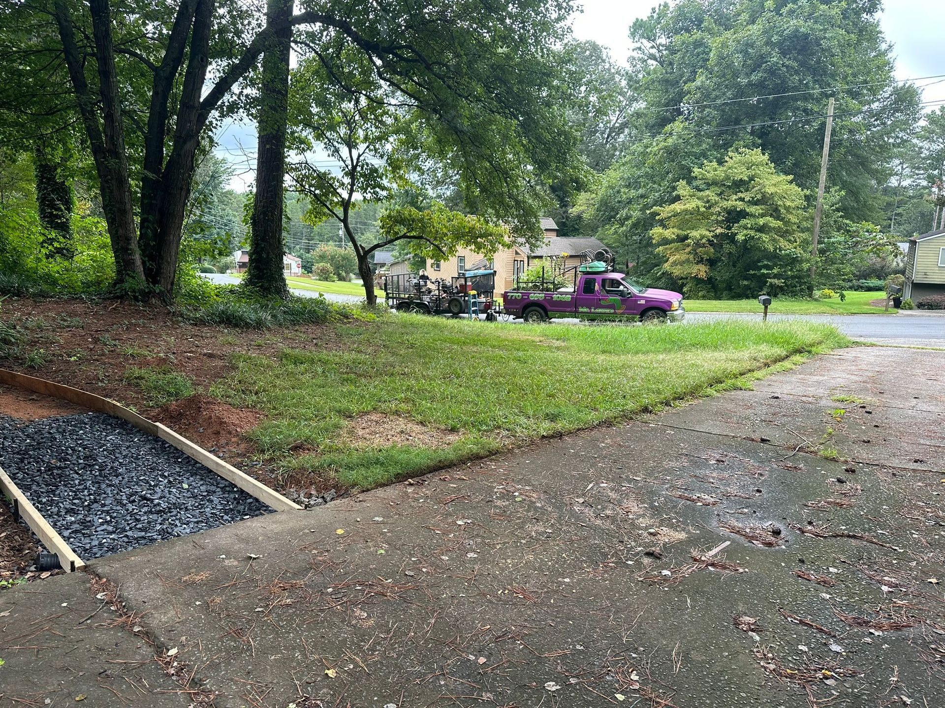Driveway with a rock-filled trough. A purple truck and trailer are parked on the grass near a house and trees.
