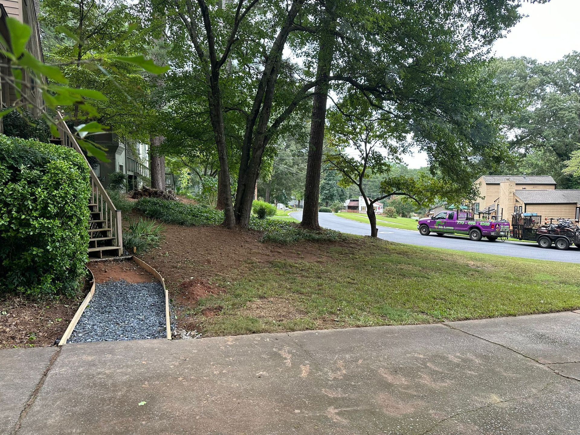 Sloping yard with grass, gravel drain, trees, and street. A vehicle is parked in the street.