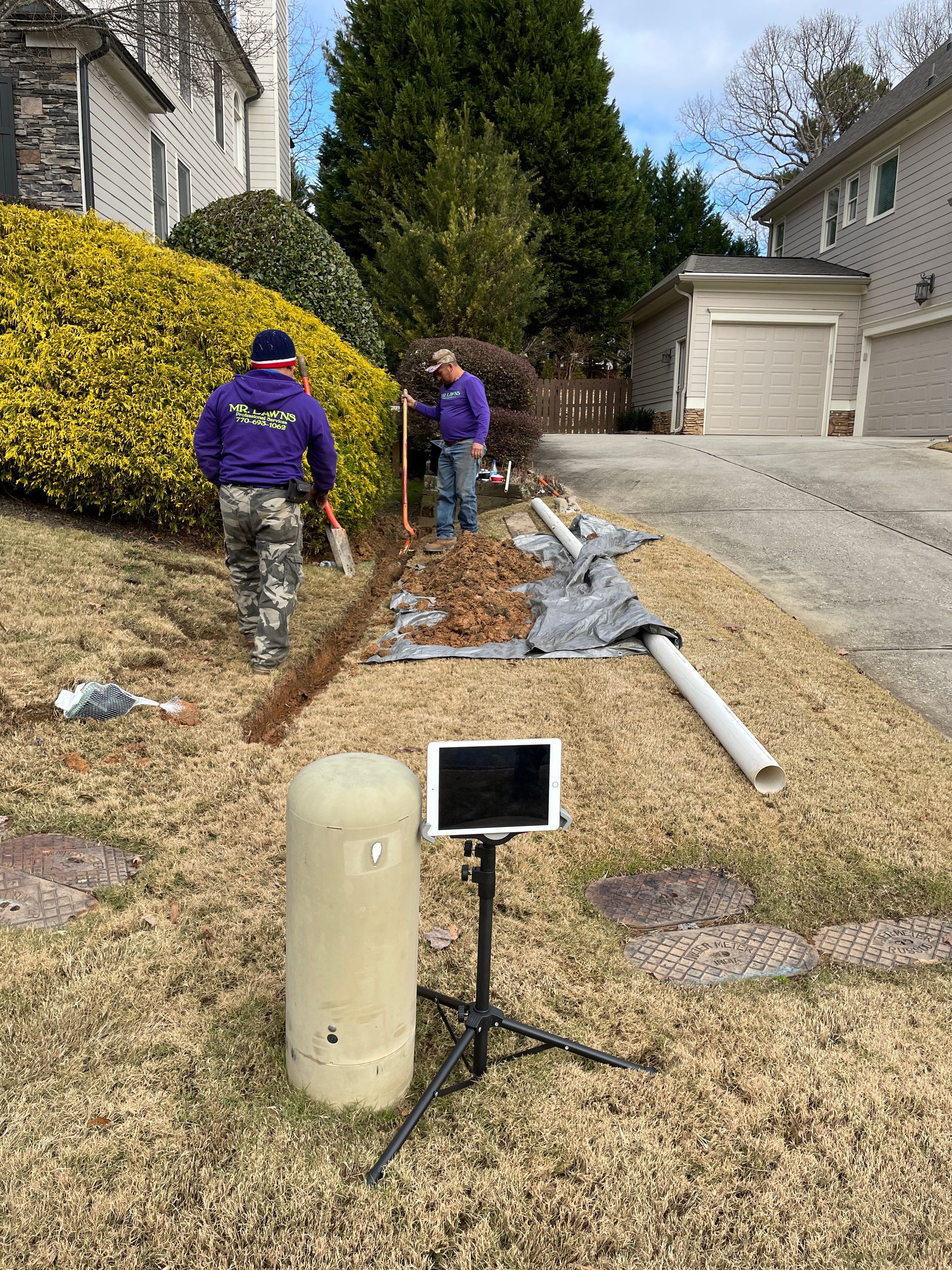 Two people installing drainage in a residential yard, with a tablet on a tripod in front.