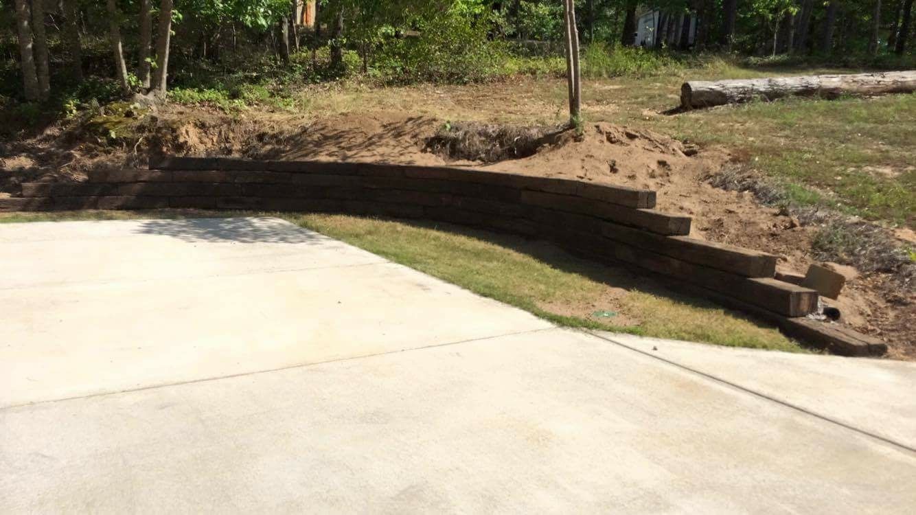 Curved retaining wall made of dark wood, with grassy area and concrete driveway in front.