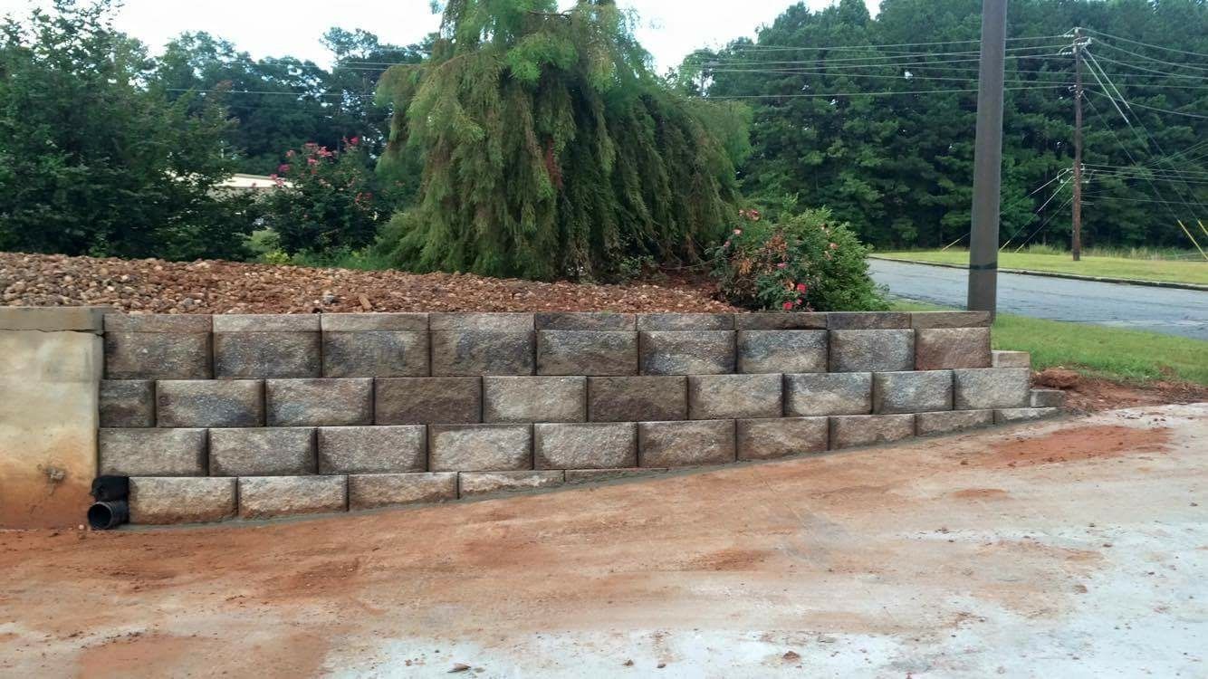 Gray retaining wall of stone blocks against a sloping landscape with trees and reddish ground.