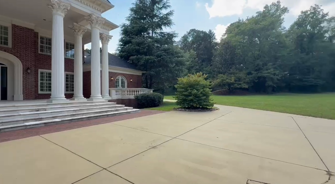 Large brick home with white columns and paved driveway, surrounded by greenery.