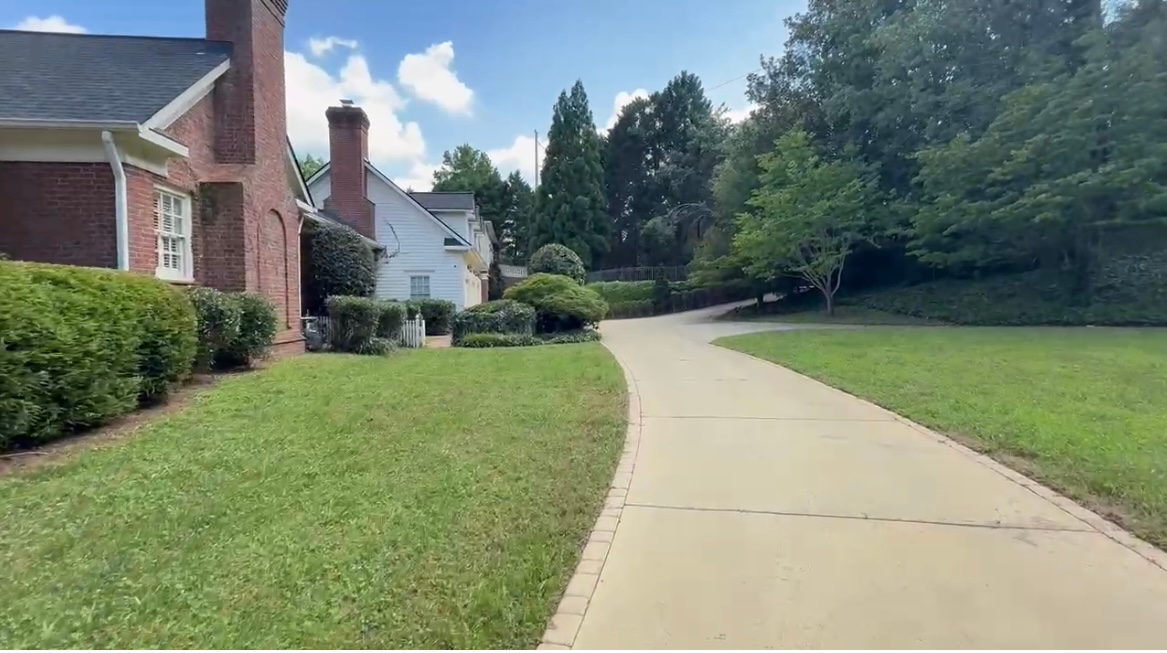 A concrete driveway curves past a grassy lawn and a brick house on a sunny day.