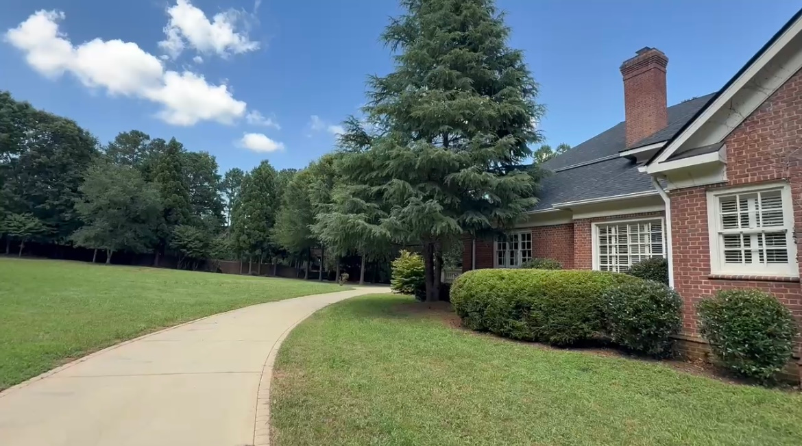Concrete path curves through green lawn toward brick house with chimney and blue sky.