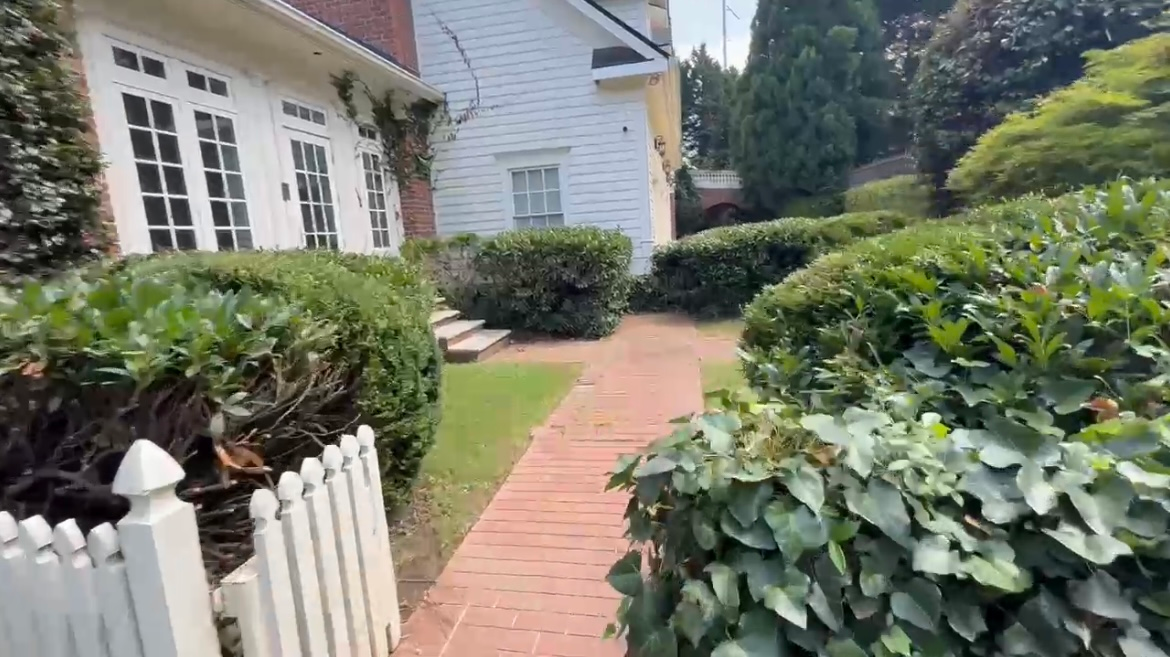 Brick pathway lined with bushes leads to a white and yellow house with a white picket fence.