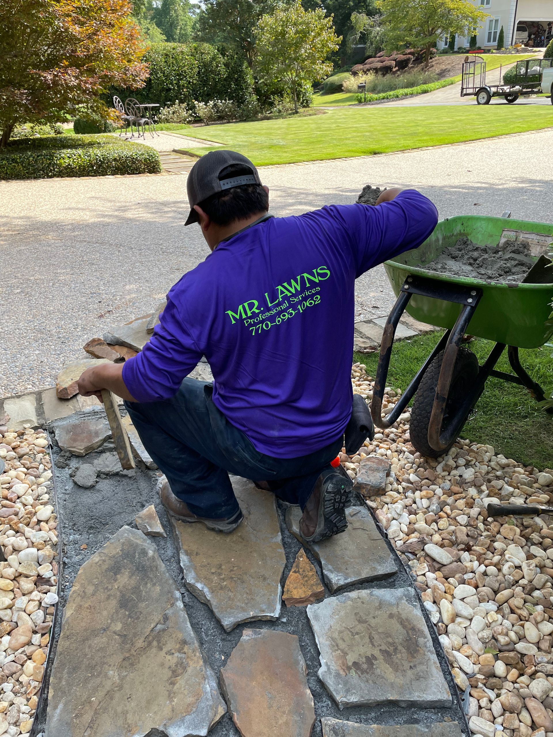 Person in purple shirt laying stone walkway. Gravel and wheelbarrow in the background.