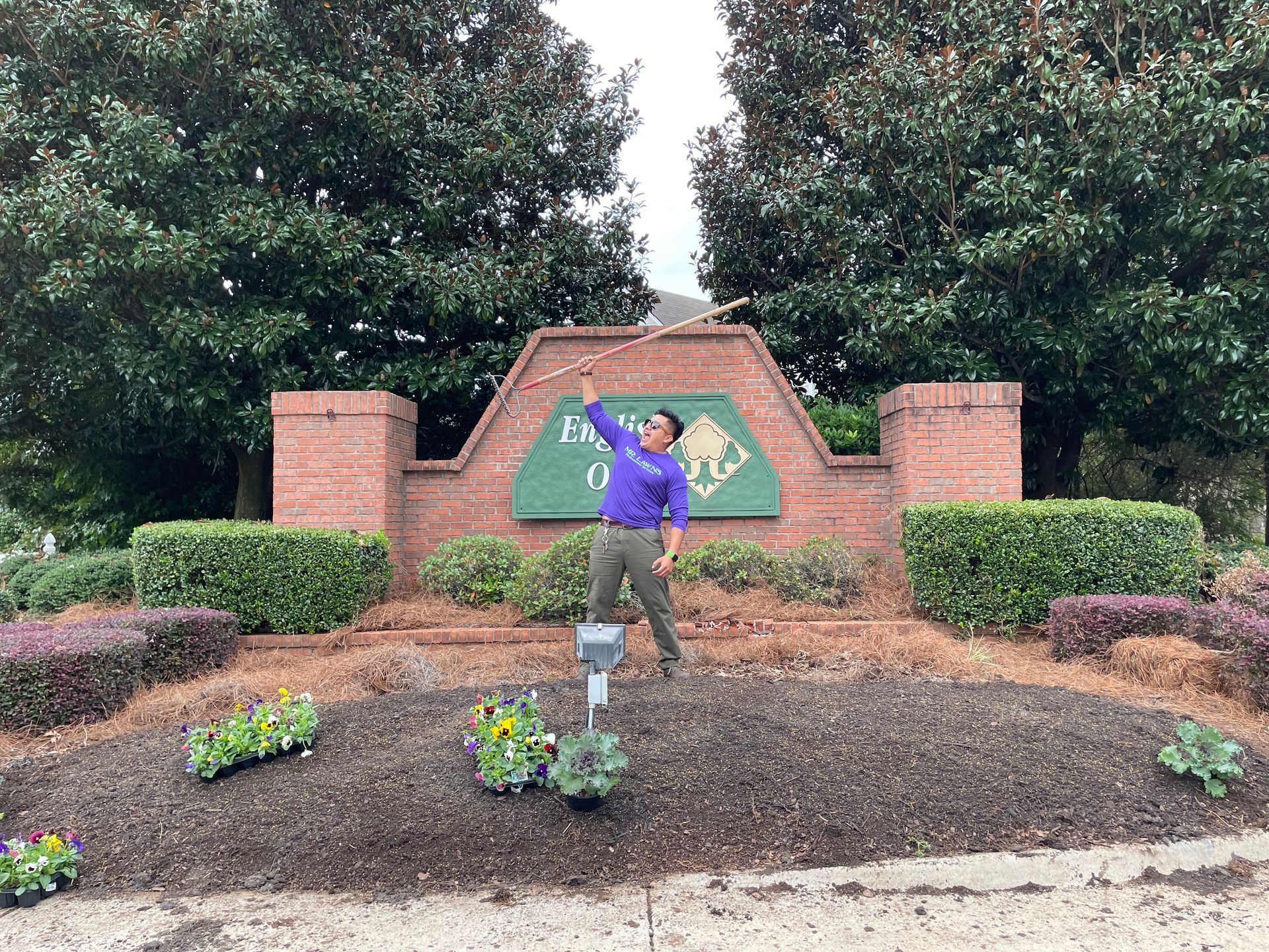 Man in purple shirt holds a stick in front of an entrance sign in a residential area.