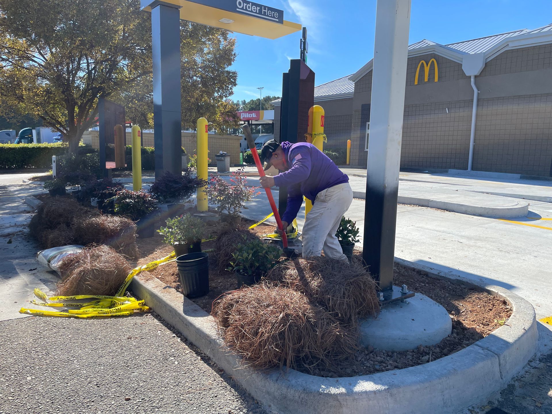 Person gardening near a McDonald's drive-thru. They are using a red tool in a flowerbed, sunny day.