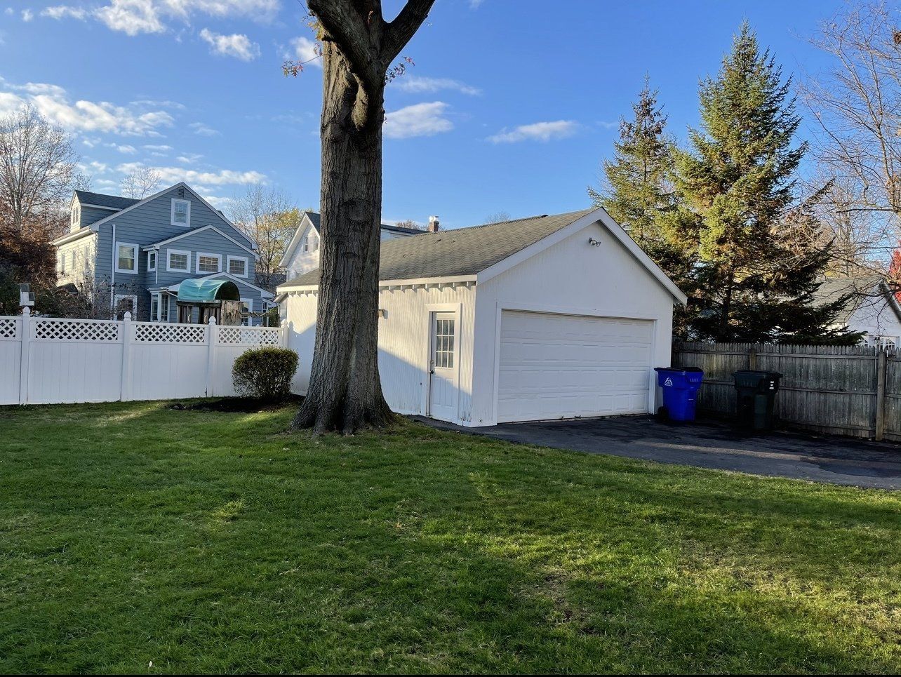 A white garage with a closed door, in front of a blue house and white fence on a sunny day.