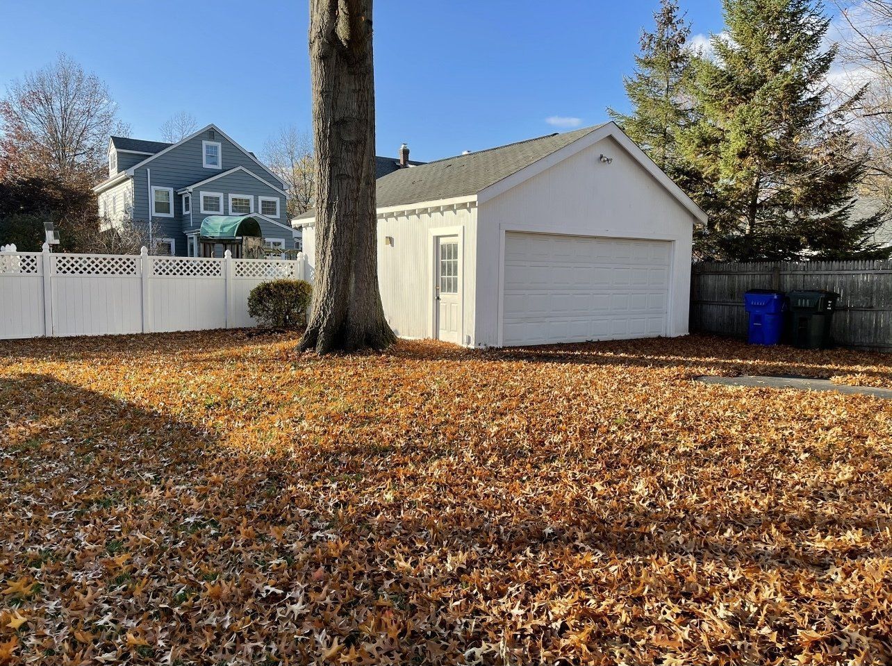 A white garage sits in a yard covered in fallen leaves, with a tree in front. A house and fence are in the background.