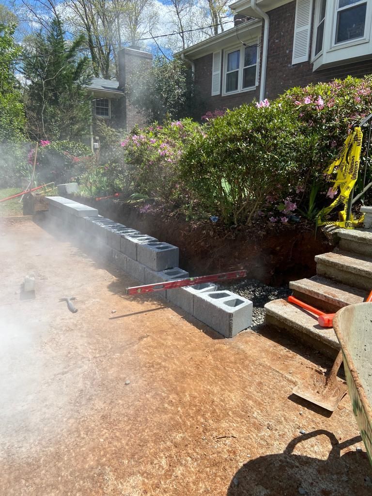 Retaining wall under construction with gray cinder blocks, near a house with azalea bushes.