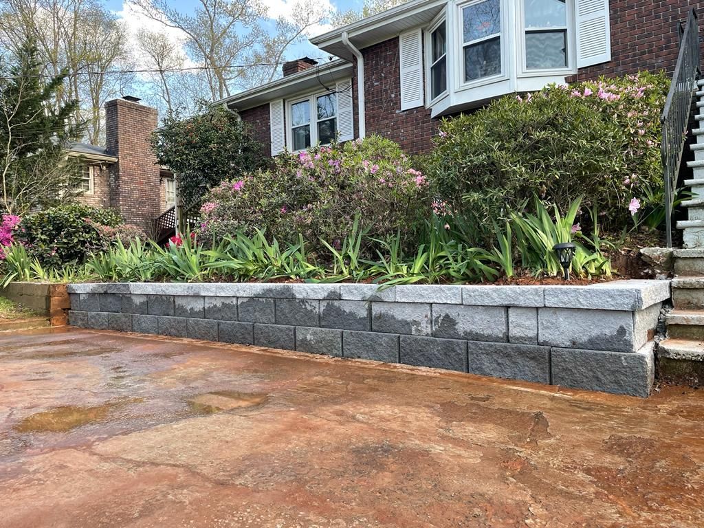 Stone retaining wall with plants in front of a brick house with white trim.