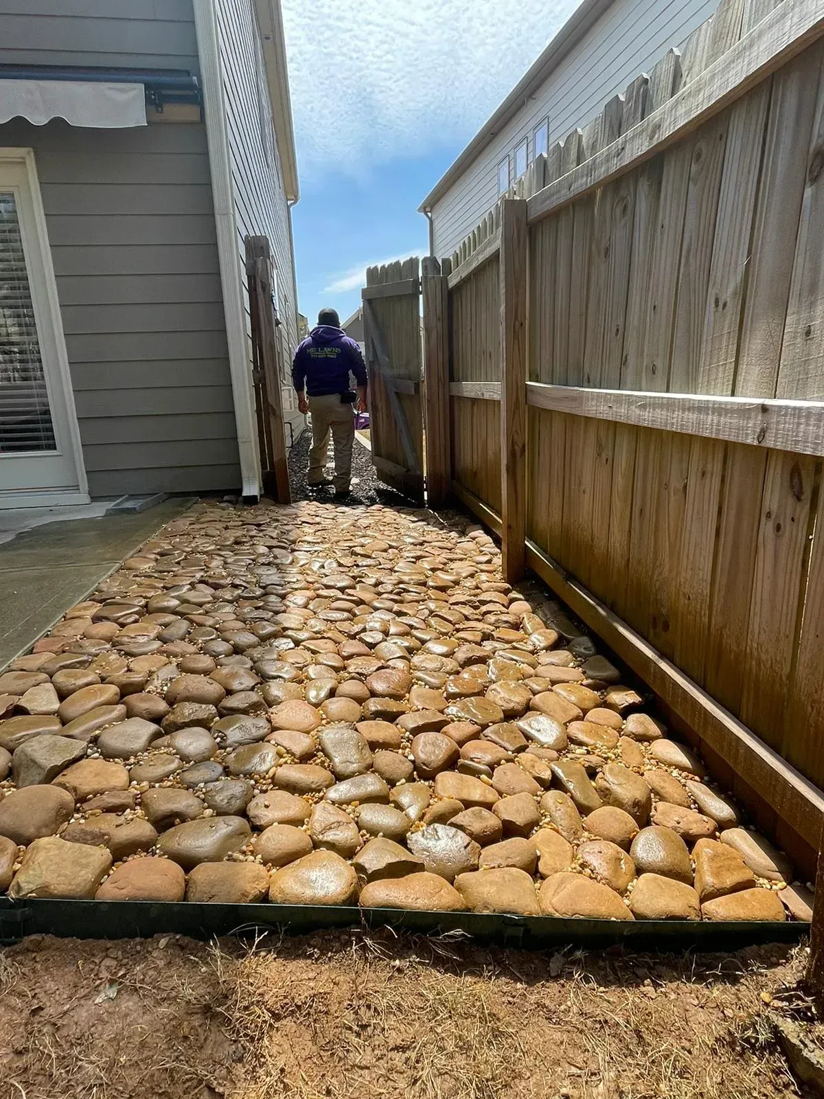 A man walks down a narrow pebble path between two wooden fences and a house.