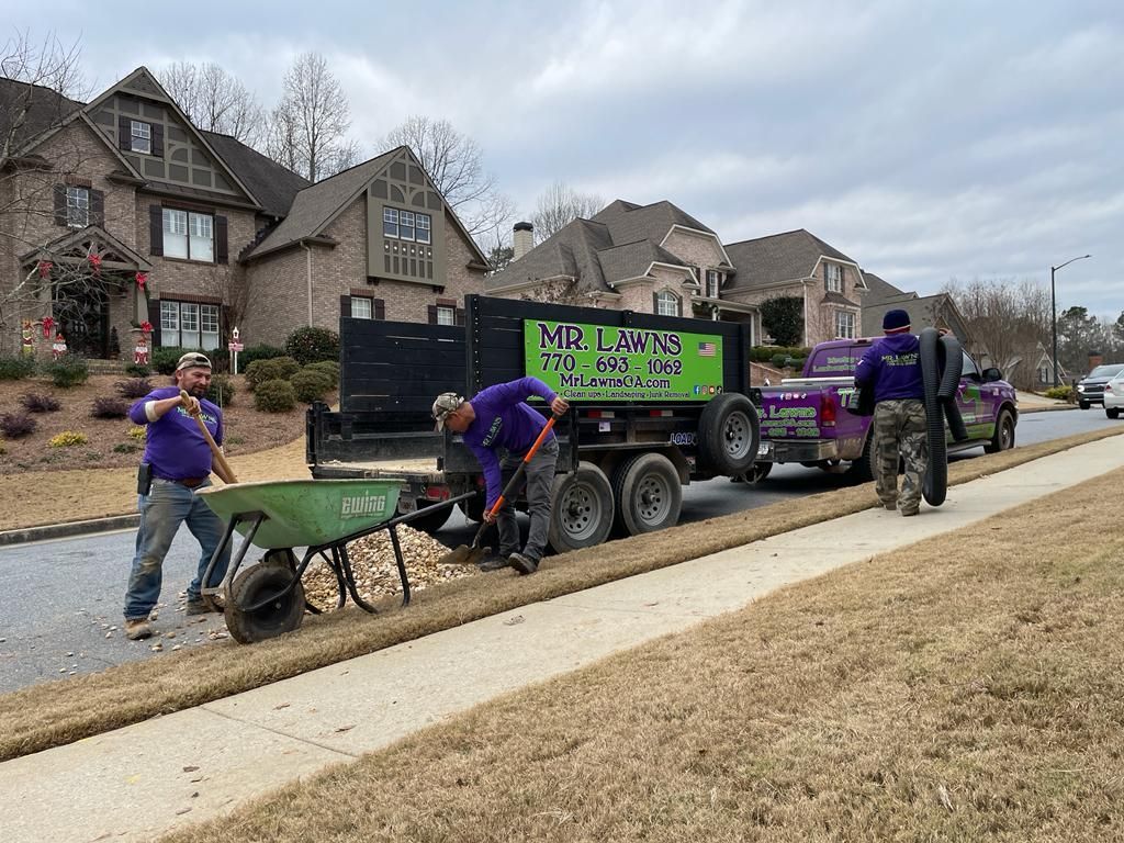 Workers in purple shirts unload rocks from a trailer along a curb in front of houses.