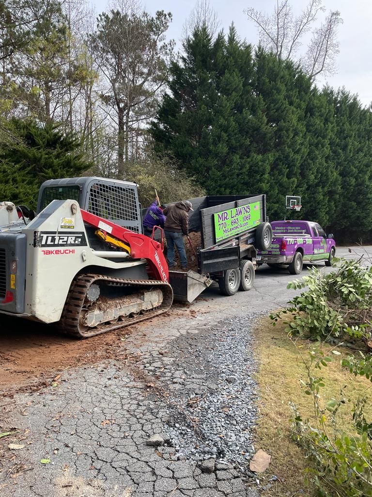 Skid steer loading a dump truck with gravel on a gravel driveway. A purple car is parked nearby.