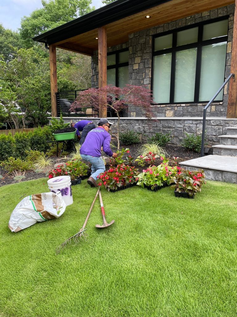People planting flowers in front of a modern house with stone facade, on a green lawn.