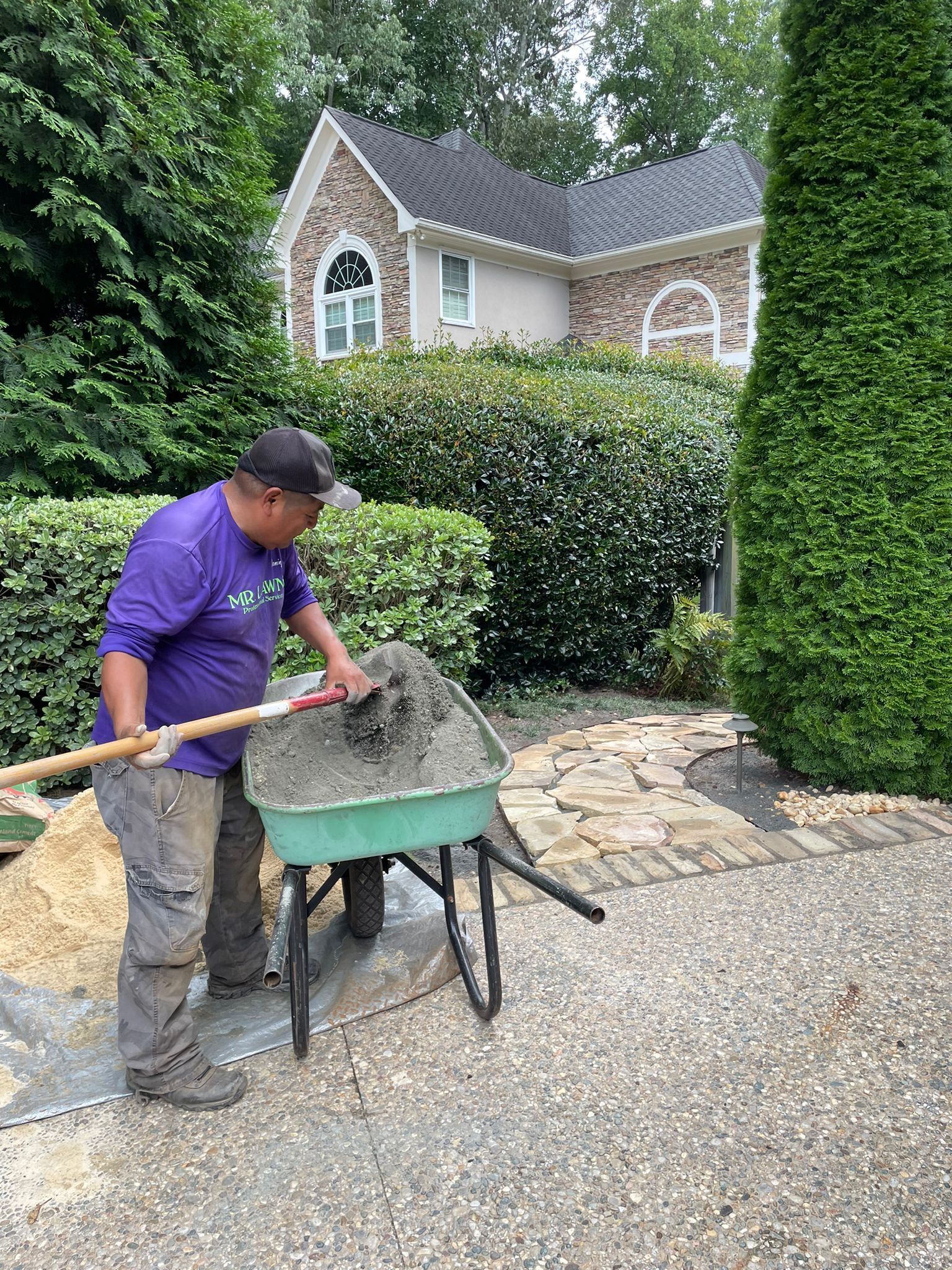 A person in purple shirt mixes cement in a wheelbarrow, with a house and landscaping in the background.