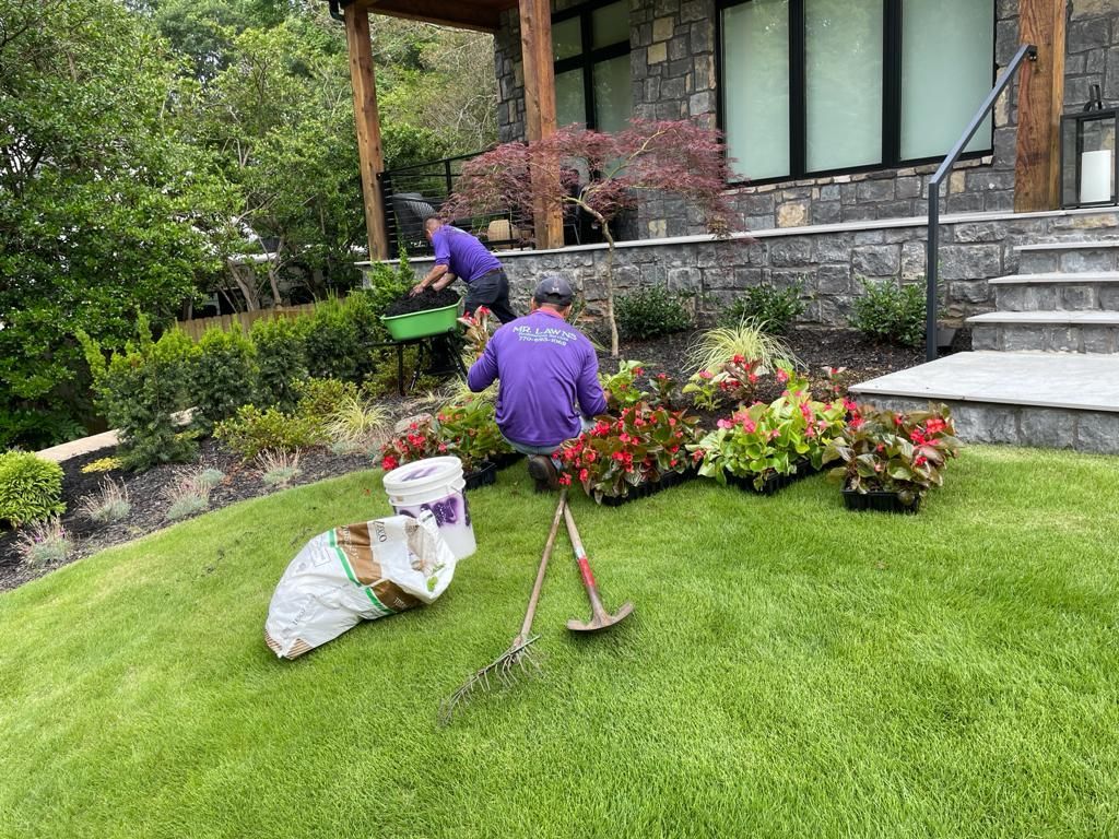 Two people planting flowers on a grassy slope next to a stone house; planting supplies are nearby.