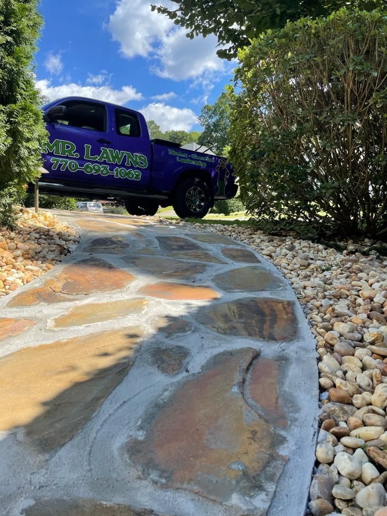 Blue truck parked on a stone pathway, landscaping in foreground, bright sky overhead.