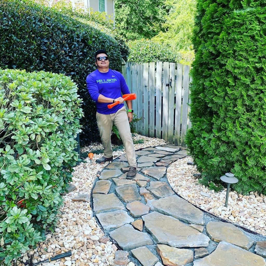 Man holding orange toy, walking on stone path in a garden, with green bushes and white fence.
