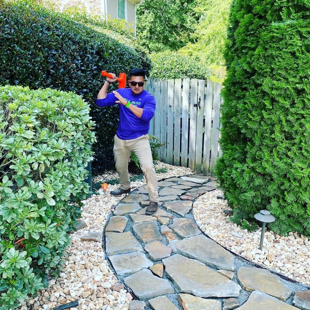 Man swings a sledgehammer on a stone path in a garden. He wears a blue shirt, khaki pants, and sunglasses.