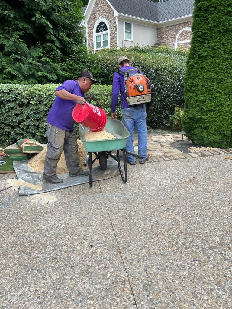 Two workers in purple shirts filling a wheelbarrow with sand, paving stones and house in background.