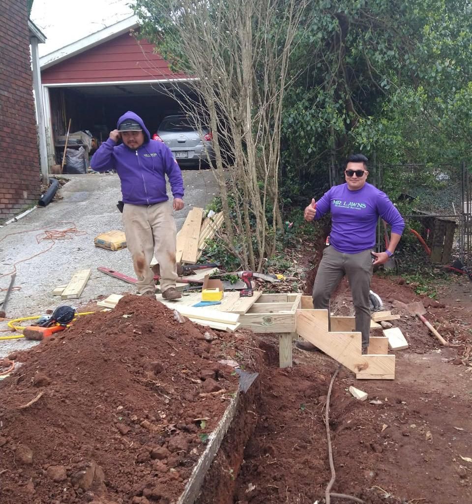 Two people in purple shirts work on landscaping project, one giving a thumbs up. Dirt pile, trench, and garage visible.