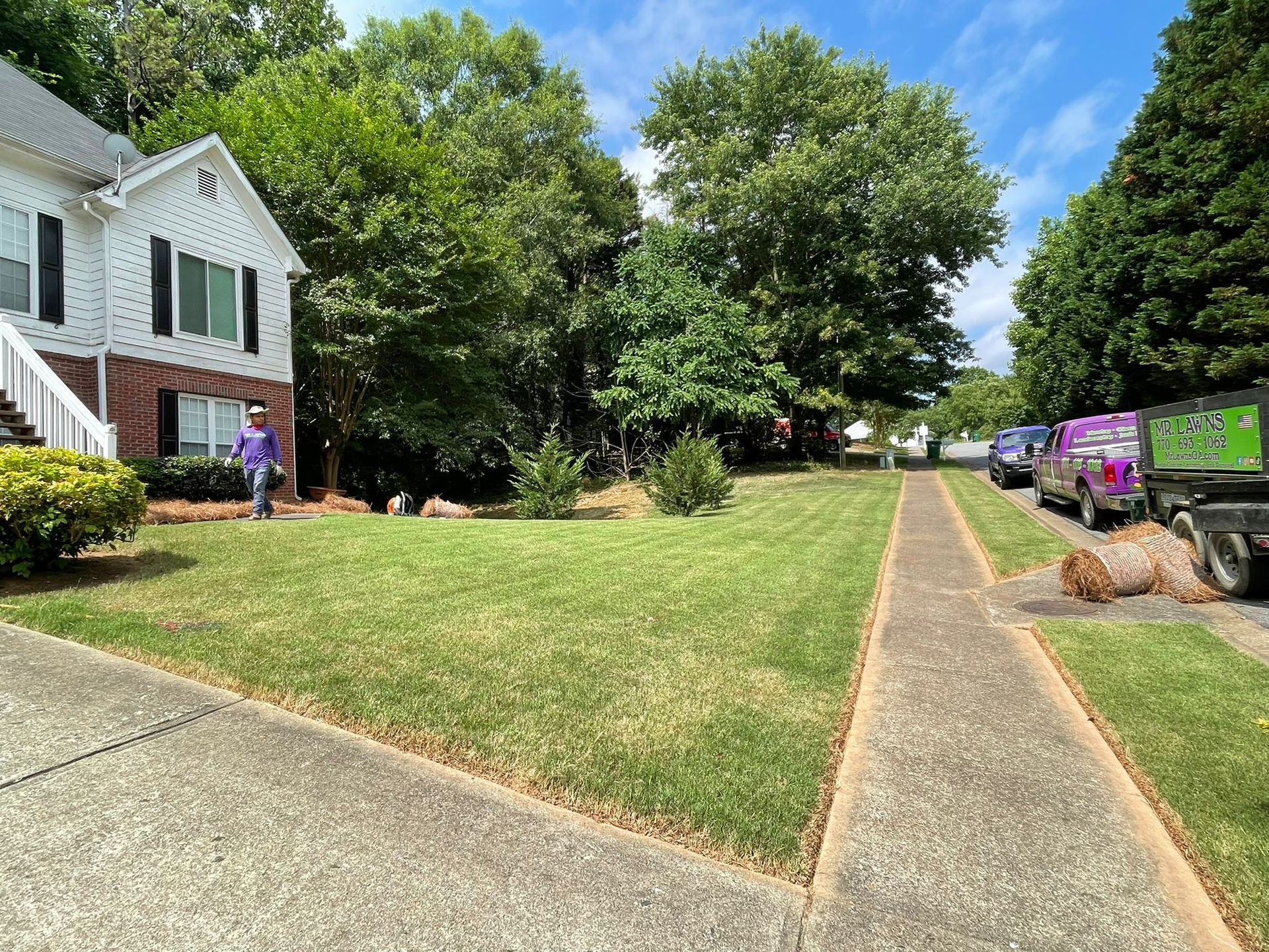 Lawn care worker edging grass near a walkway. White house, green trees, and parked trucks.
