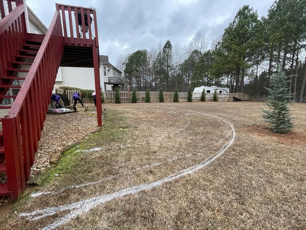Backyard scene: a deck with stairs, curved white lines in the grass, and workers. Trees and a camper are in the background.