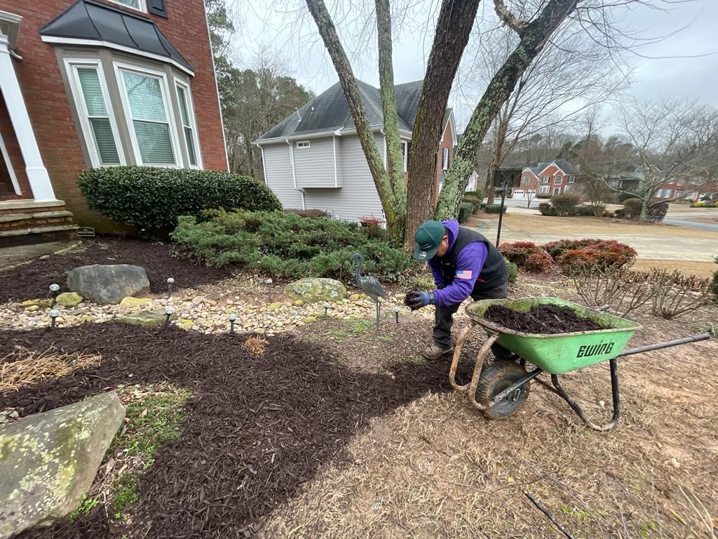 Person spreading mulch in a landscaped yard next to a brick house. A wheelbarrow is nearby.