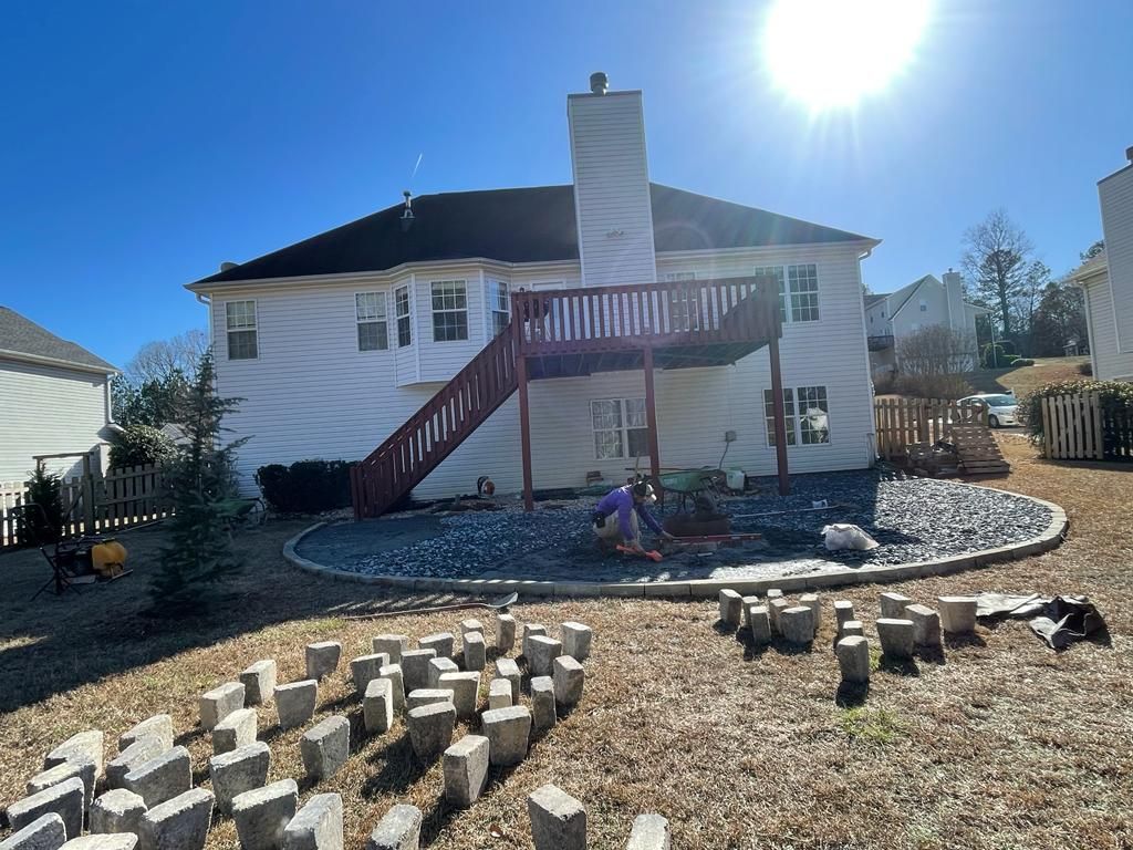 Backyard with two-story house, wooden deck, chimney, and landscaping being worked on by two people. Sunny day.
